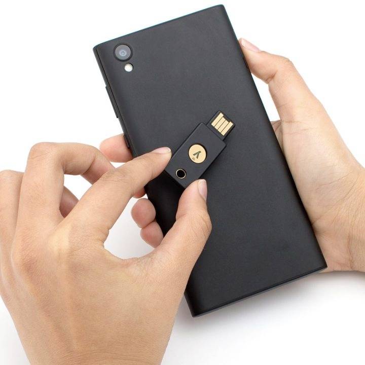 Osoba drží smartphone s připojeným bezpečnostním USB klíčem. USB klíč má své logo a slouží k autentizaci.