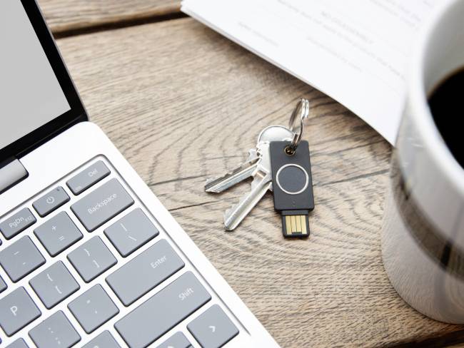 Klávesnice notebooku se dvěma kovovými klíči a bezpečnostním USB klíčem na dřevěném stole, vedle šálek kávy a dokument.