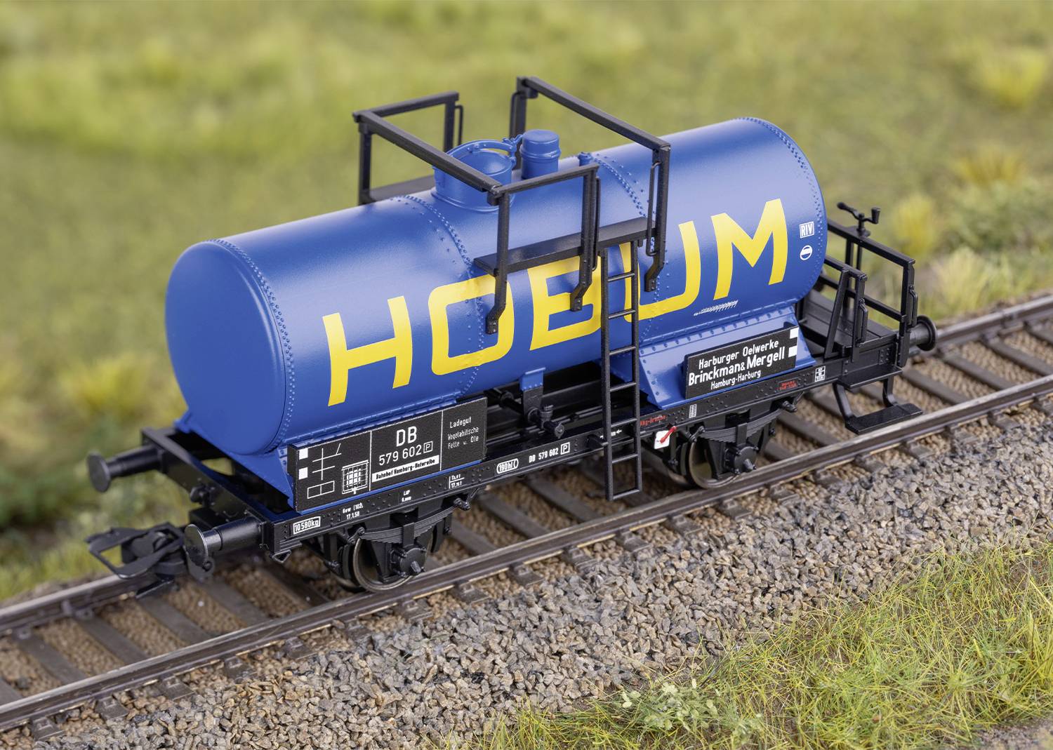 Modrý cisternový vůz na kolejích modelové železnice. Na nádrži je žlutým písmem napsán nápis 'HOEBUM'.