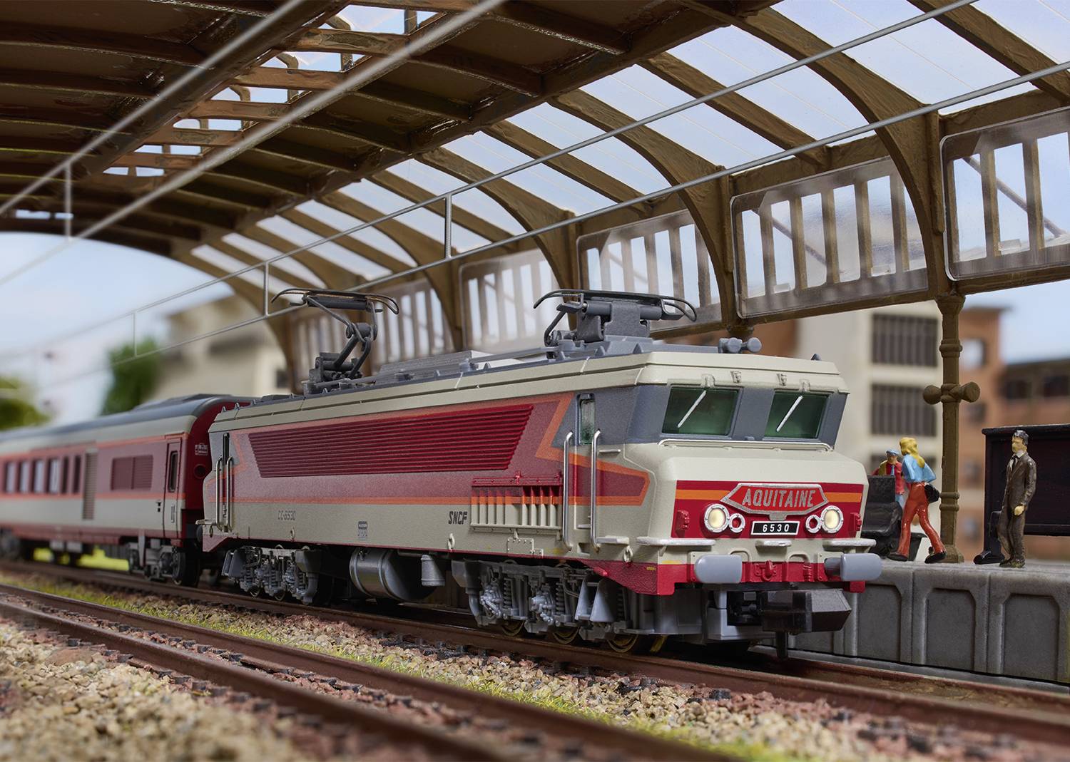Model elektrické železnice s červeným a šedým vlakem jede pod nádražní střechou. Lidé stojí na nástupišti.