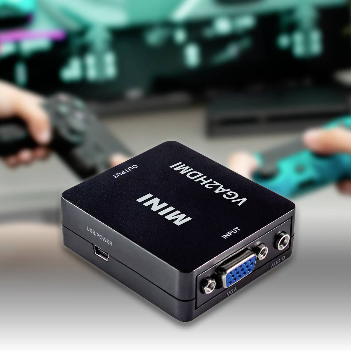 Před vámi leží konverterbox 'Mini VGA2HDMI'. V pozadí jsou rozmazaně vidět dvě ruce s herními ovladači.