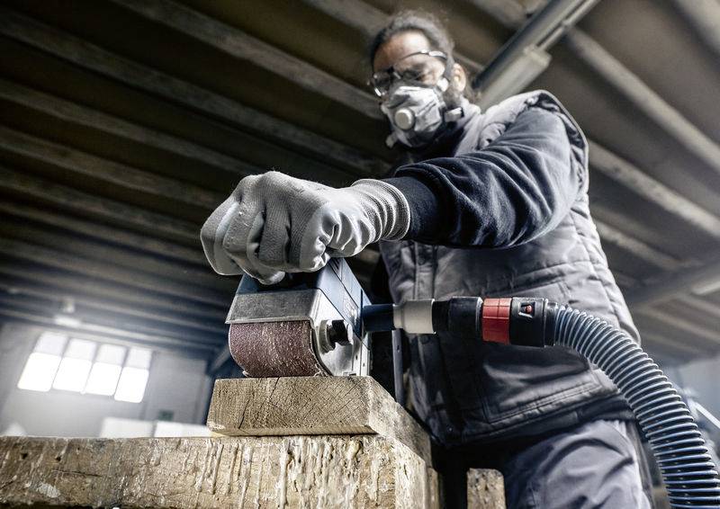 Osoba brousí dřevěný díl elektrickým brusným zařízením. Má na sobě ochrannou masku a rukavice v prostředí dílny.