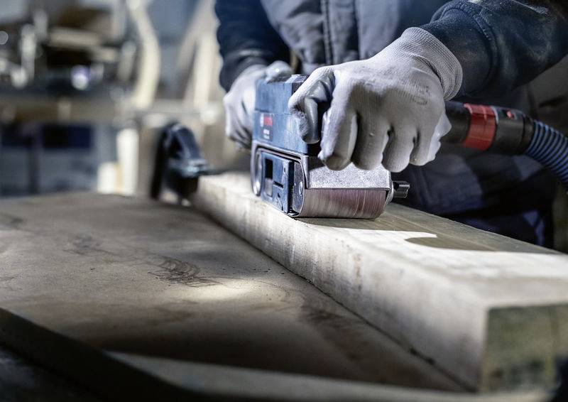 Osoba brousí dřevěnou desku v dílně. Ruce jsou oblečeny do šedých pracovních rukavic a obsluhují elektrické brusné zařízení.