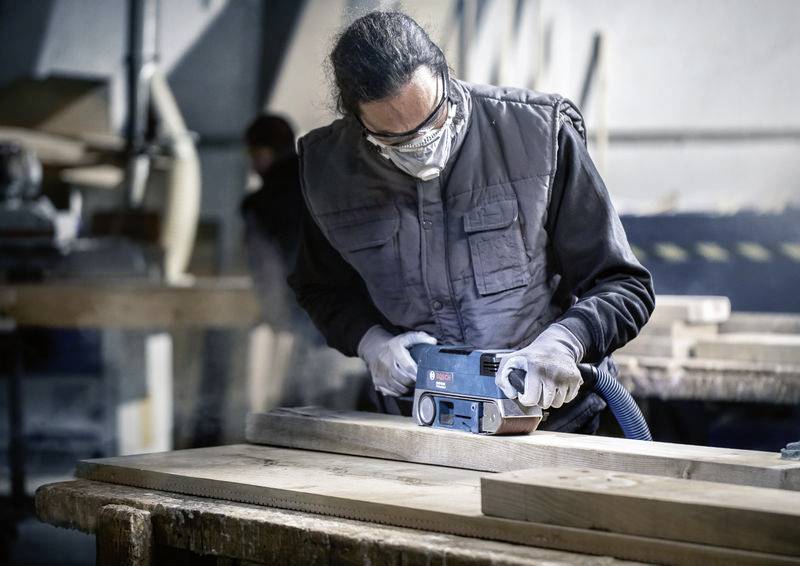 Osoba brousí dřevěnou desku bruskou ve stolařské dílně. Má nasazené ochranné brýle a respirátor.