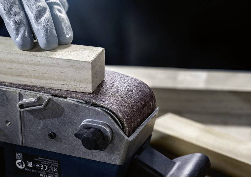 Stroj obrušuje dřevěnou desku. Rukavicí chráněná ruka drží desku pevně a vede ji přes brusný pás.