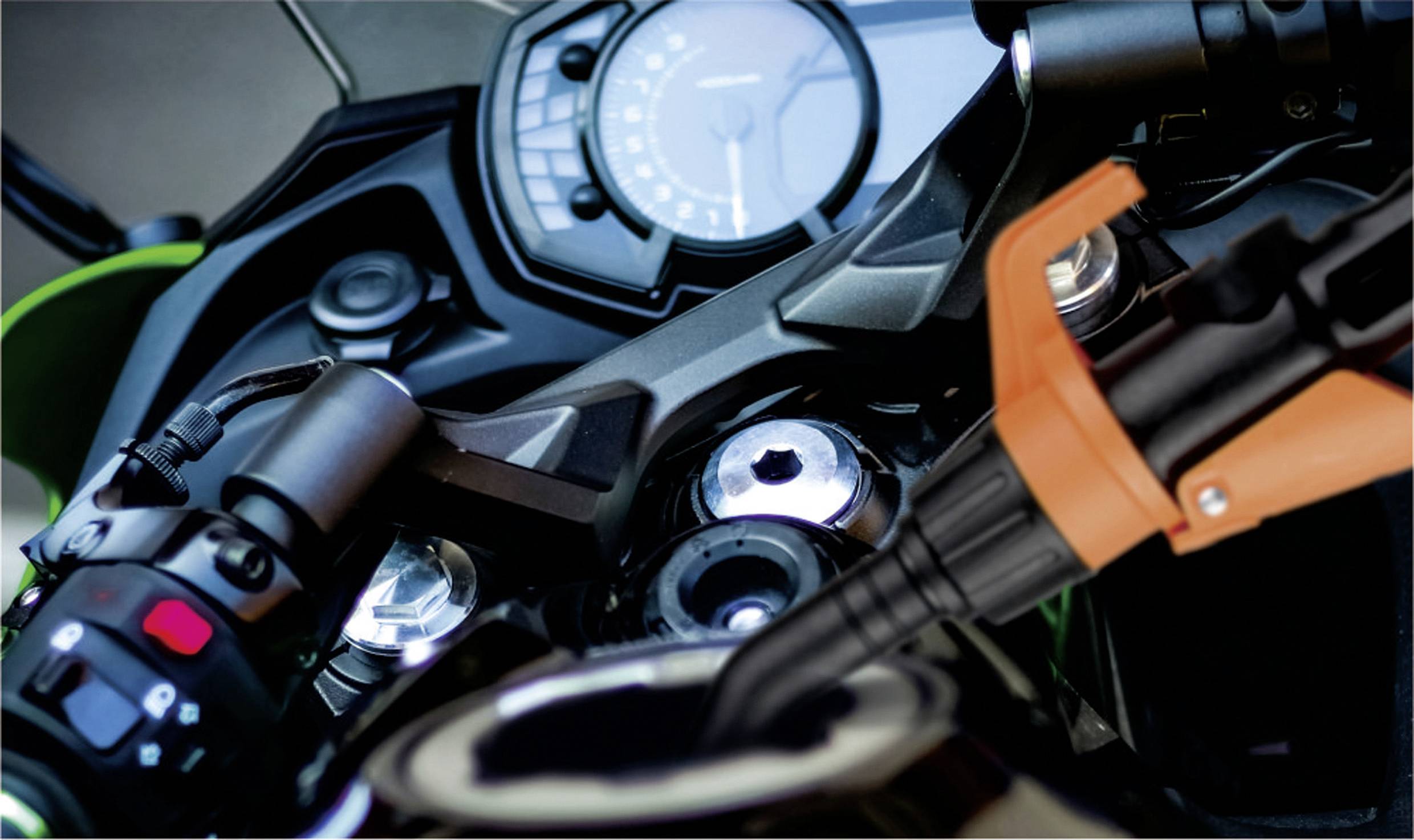 Detailní záběr motocyklového kokpitu s tachometrem, oranžovou brzdovou pákou a ovládacími tlačítky na řídítkách. Zaměřeno na rychlost a kontrolu.