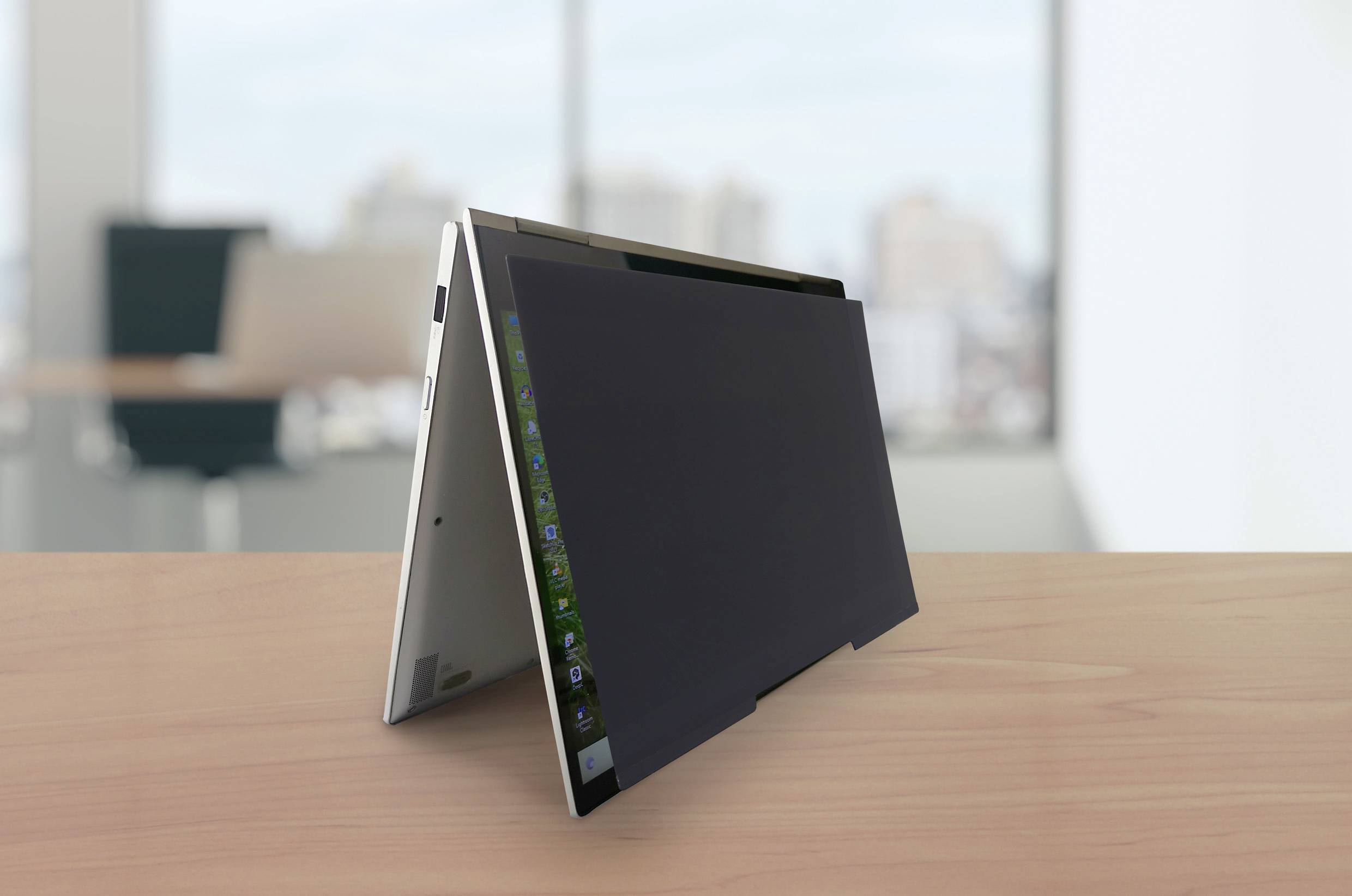 Konvertibilní notebook postavený v módu stanu na stole před světlým kancelářským pozadím.