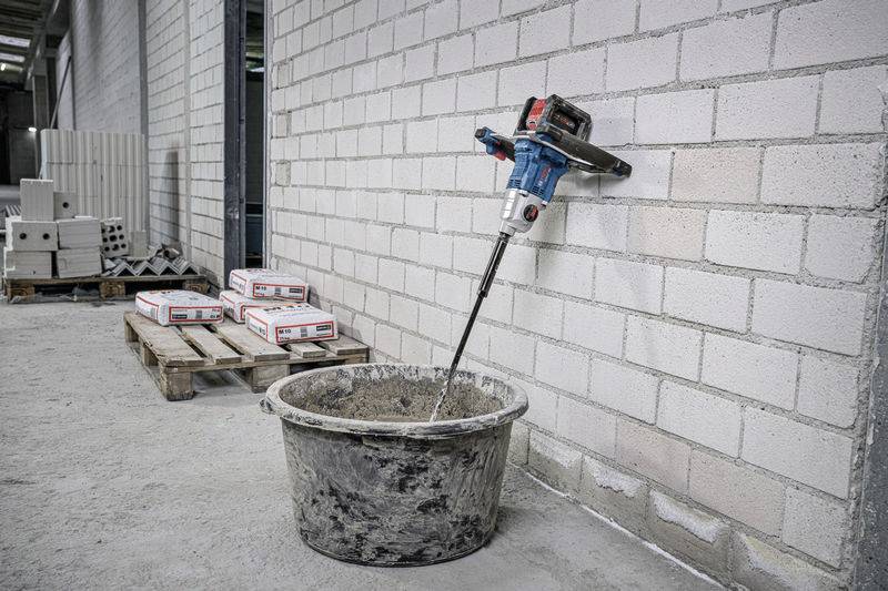 Kbelík barvy je míchán elektrickým míchadlem ve velké plastové nádobě v rámci stavebního projektu. V pozadí jsou na paletách naskládány cihly.