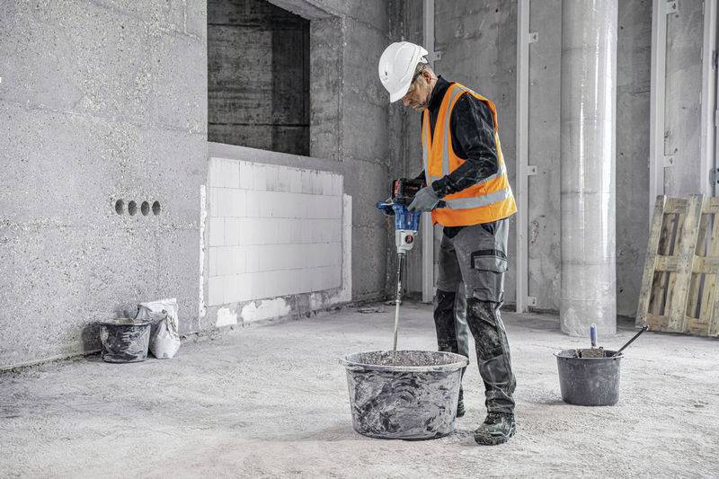 Dělník míchá materiál elektrickým míchadlem ve velkém kbelíku v betonové místnosti. Je oblečen v bezpečnostním oděvu.