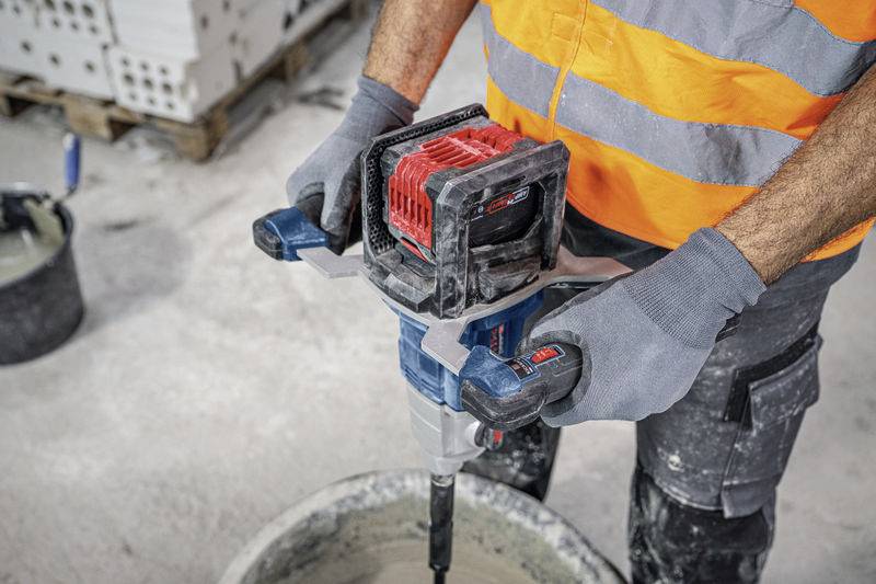 Osoba míchá cement modrým a červeným elektrickým míchadlem. Je oblečena v bezpečnostním oblečení, včetně rukavic a vesty.