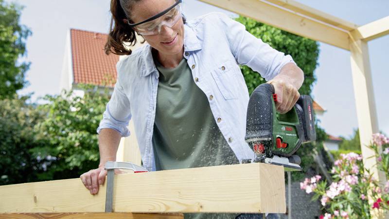 Osoba řeže zelenou elektrickou přímočarou pilou dřevěnou desku. Má ochranné brýle a nachází se venku.