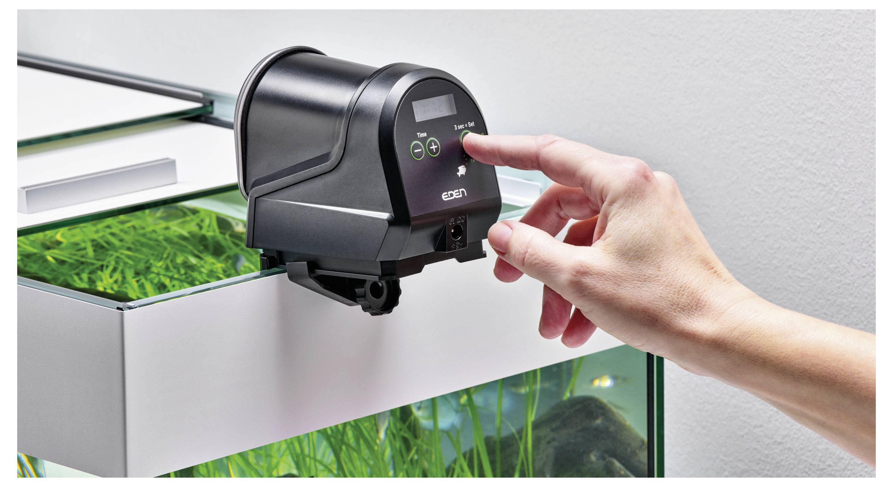 Osoba nastavuje digitální časovač na automatickém krmítku pro ryby, které je připevněno na okraj akvária s zelenými rostlinami uvnitř.