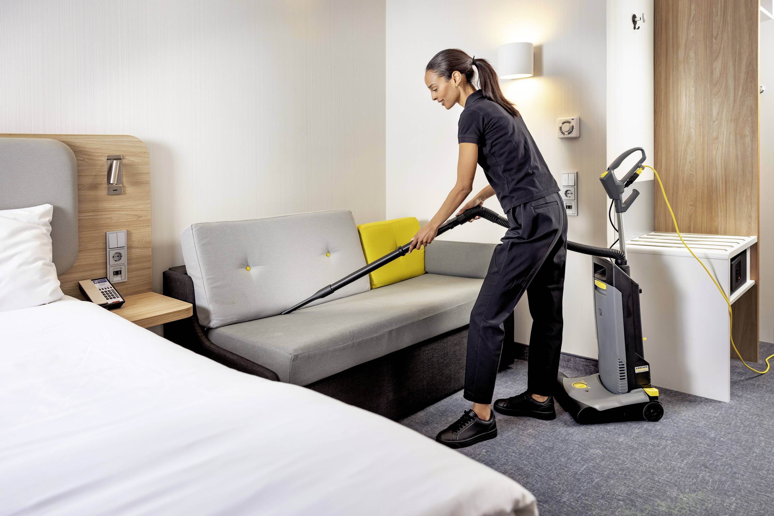 Osoba vysává pohovku v hotelovém pokoji. V popředí je vidět postel.