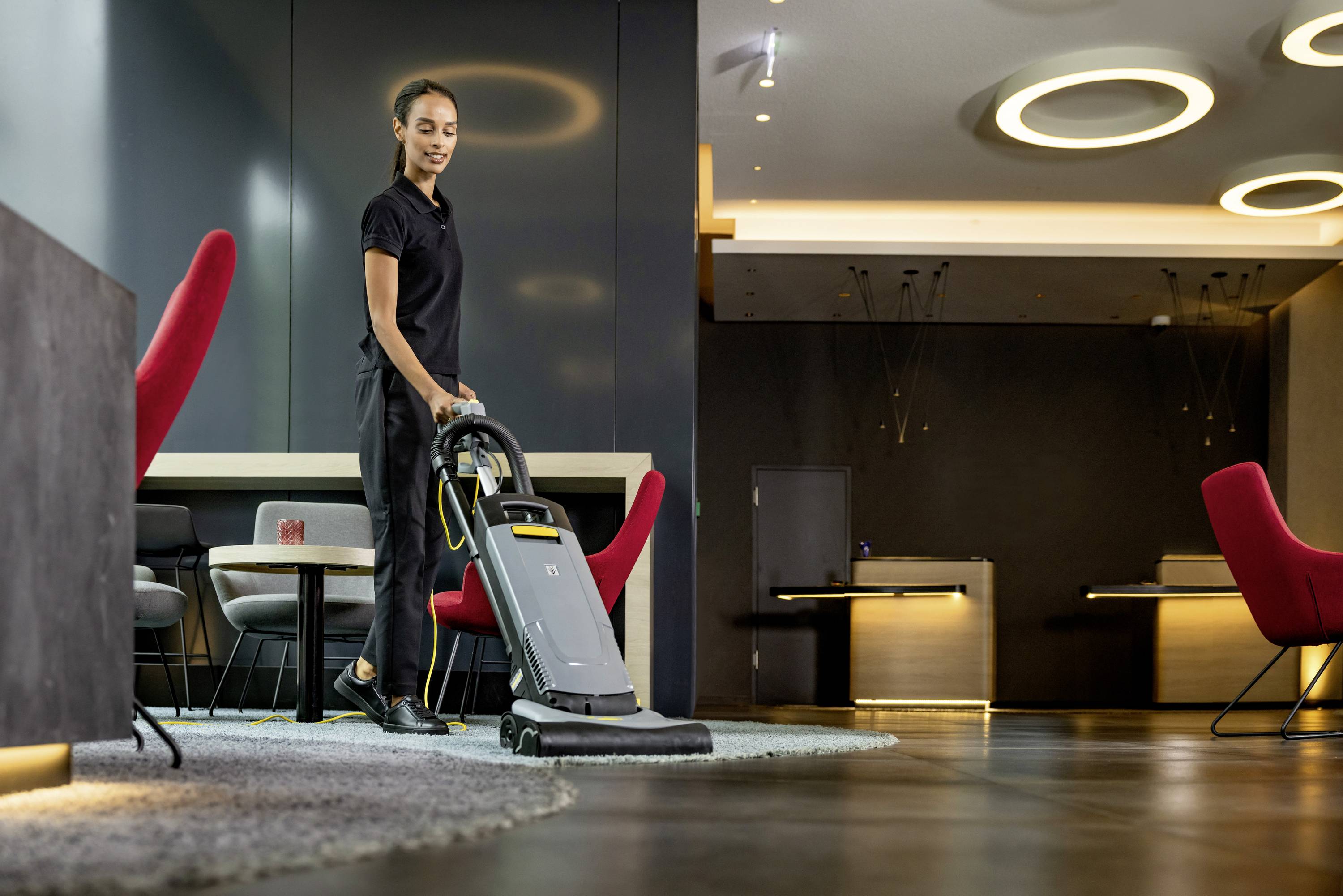 Osoba vysává moderní místnost s červenými židlemi a stolem na velkém koberci.