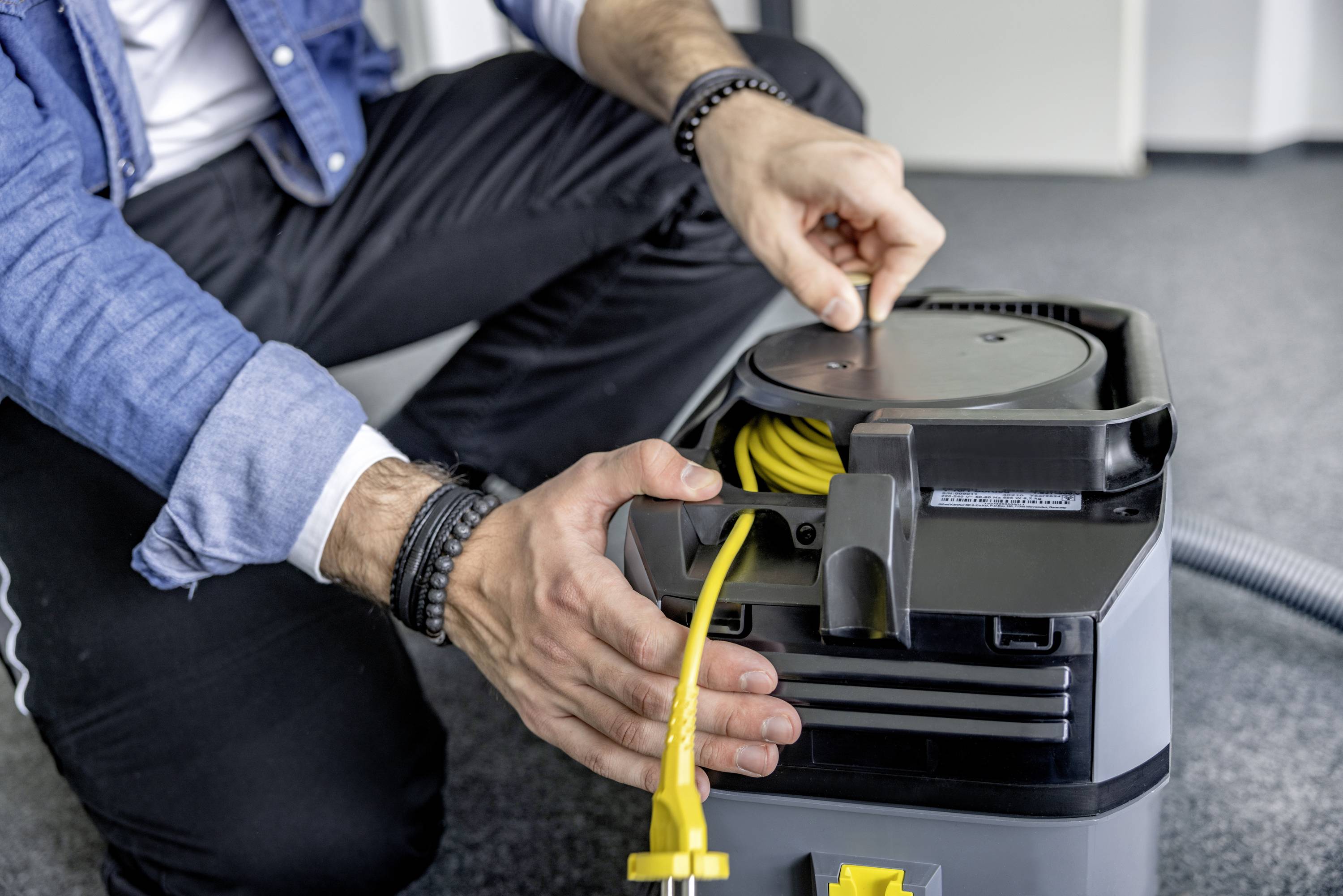 Osoba navíjí žluté elektrické napájecí vedení šedého vysavače, zatímco klečí. Ruce jsou soustředěné.