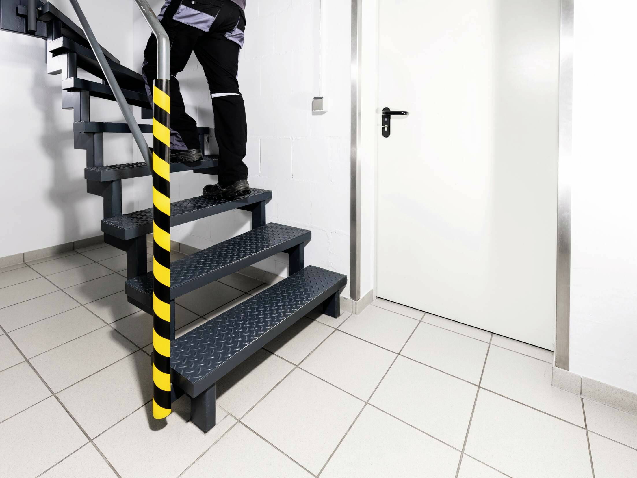 Osoba stoupá po kovových schodech, které jsou zajištěny bezpečnostním sloupkem s žluto-černým pruháním.