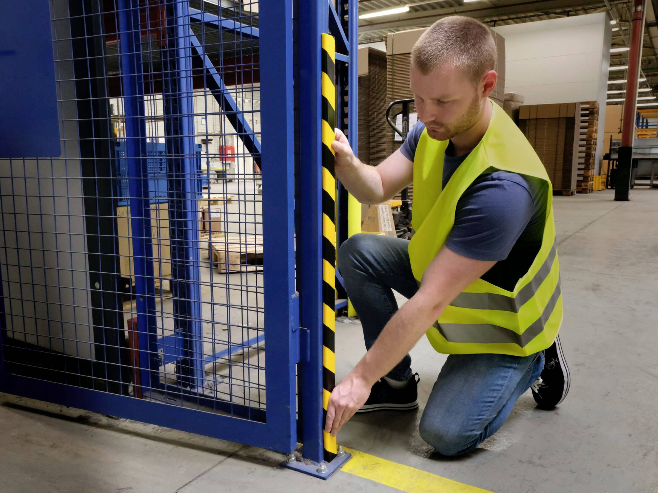 Muž ve výstražné vestě kontroluje bezpečnostní zámek skladového prostoru. Klečí a prohlíží stabilní kovovou konstrukci.