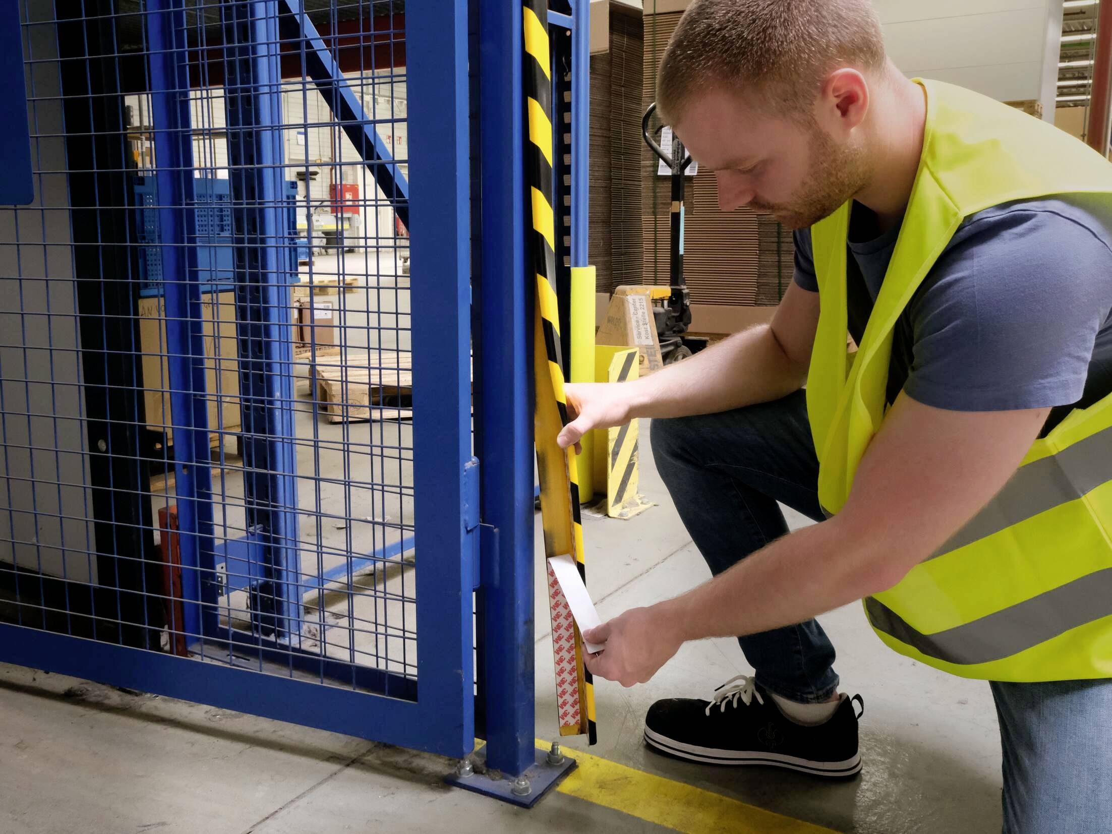Muž v bezpečnostní vestě a džínách kontroluje bezpečnostní pásku na kovovém vratech v průmyslovém prostředí.