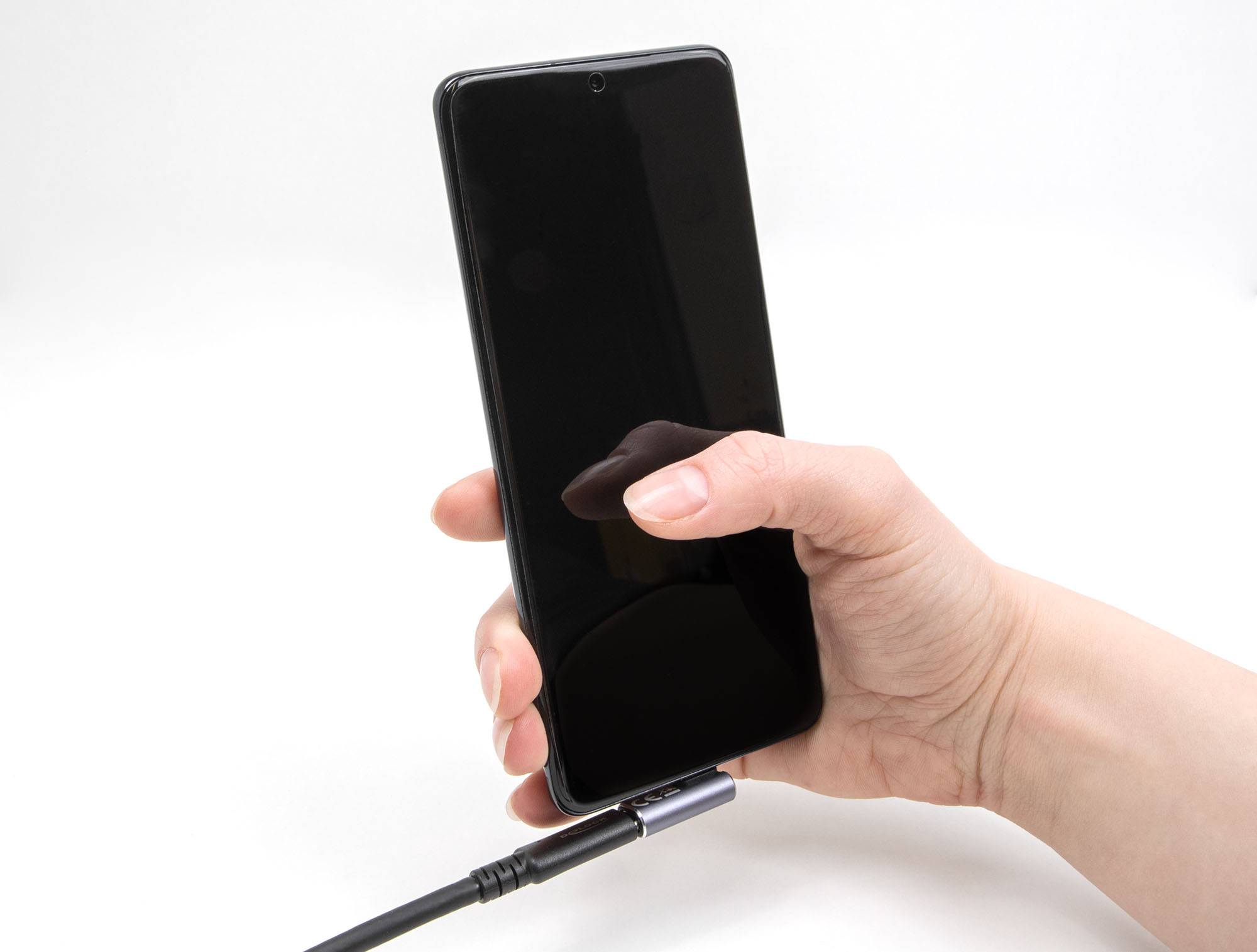 Jedna ruka drží smartphone připojený k nabíjecímu kabelu. Displej je černý a zařízení není zapnuté.
