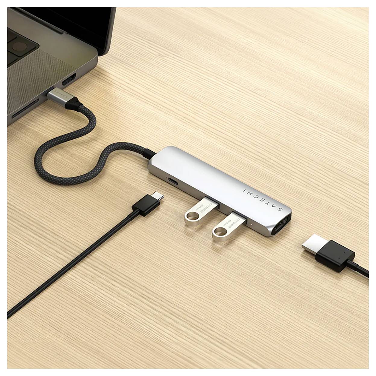 USB-C hub je připojen k laptopému počítači. Dva USB flash disky a kabel HDMI jsou připojeny k hubu. Hub leží na dřevěném stole.