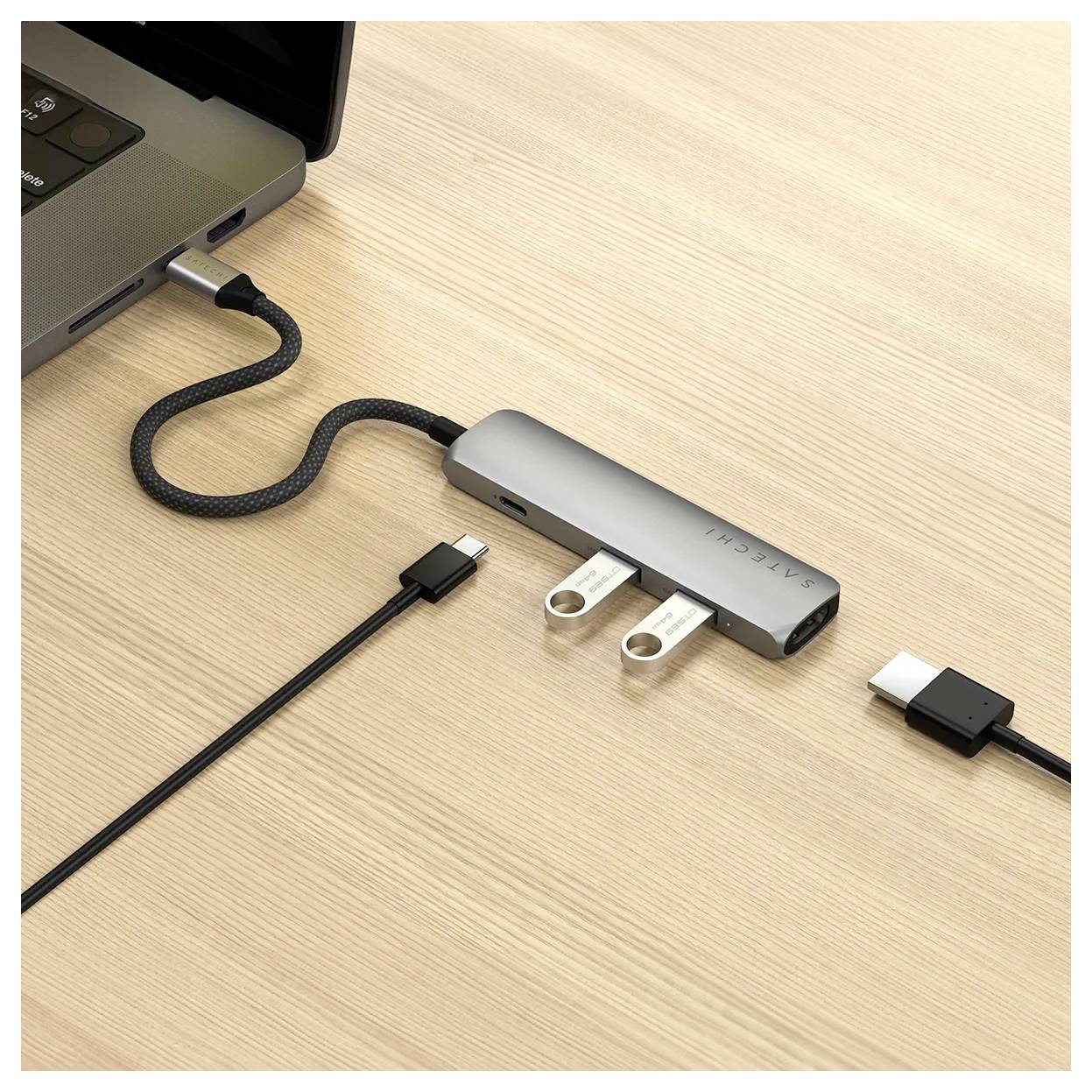 Notebook je připojen přes USB hub s několika porty. Na dřevěném stole jsou viditelné USB a HDMI kabely.
