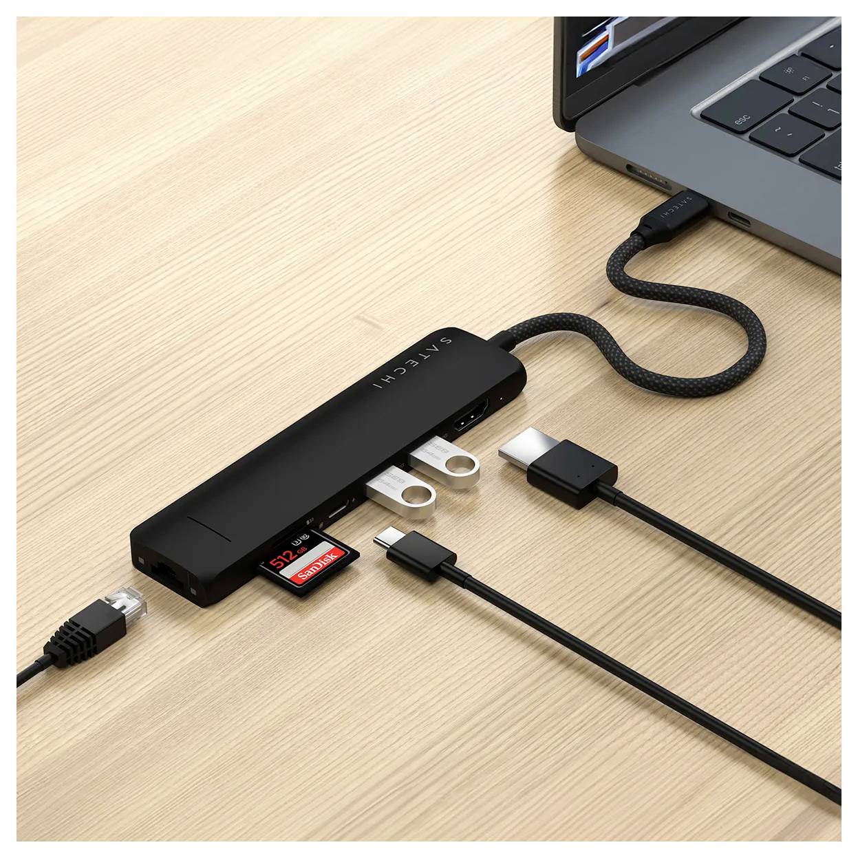 Černý USB-C hub s několika porty položený na dřevěném stole, připojený k notebooku. Jsou viditelné kabely a slot na SD kartu.