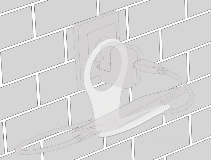 Telefon se nabíjí v zásuvce pomocí držáku, který zařízení podpírá a vede kabel. V pozadí je vidět bílá obložená zeď.