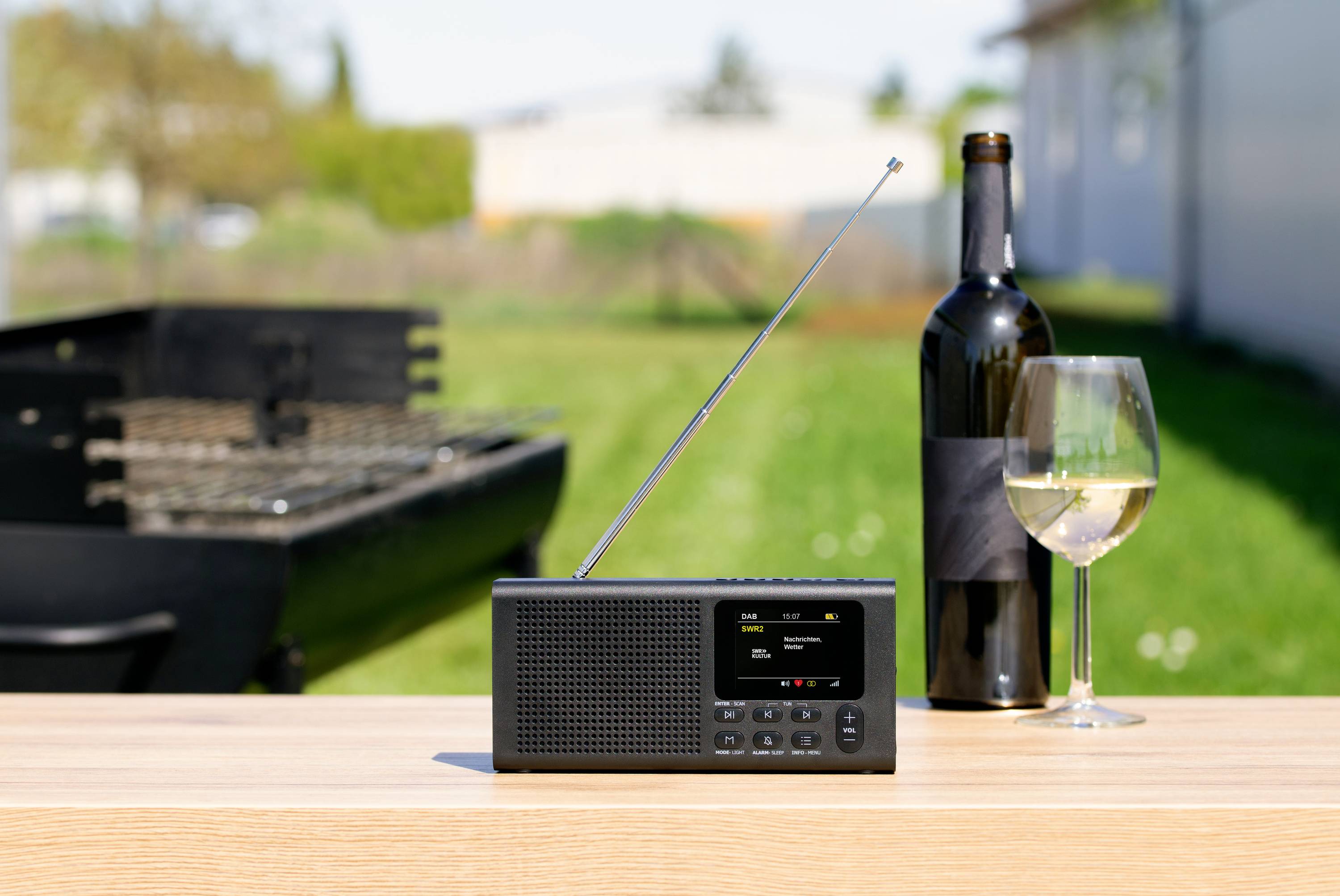 Rádio s vysunutou anténou stojí na stole vedle láhve vína a sklenice bílého vína venku, v pozadí gril.