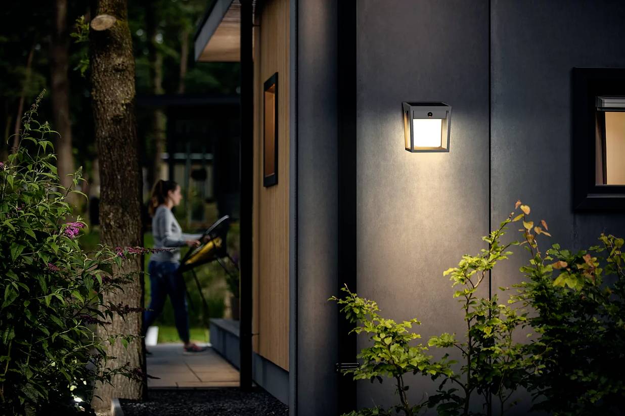 Osoba stojí venku za soumraku před moderní budovou. Venkovní osvětlení je zapnuté.