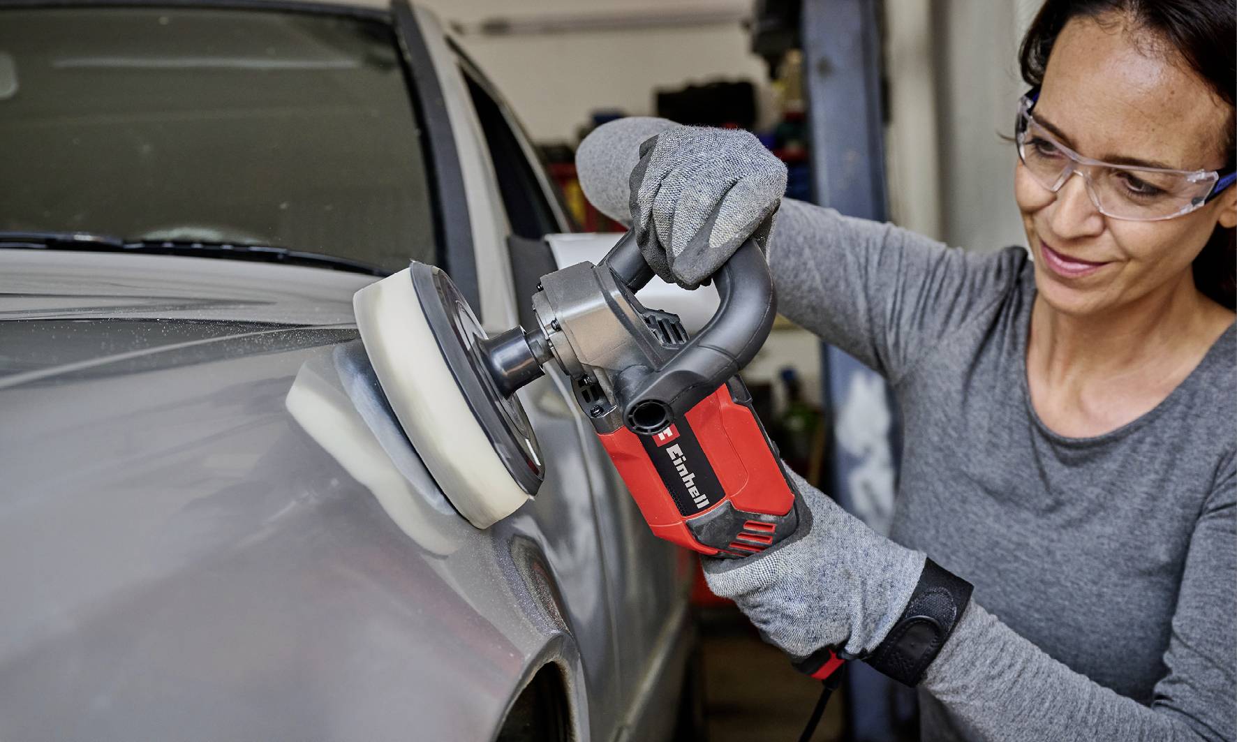 Osoba leští horní část šedého automobilu pomocí elektrické leštičky, přičemž má nasazené ochranné brýle a rukavice.