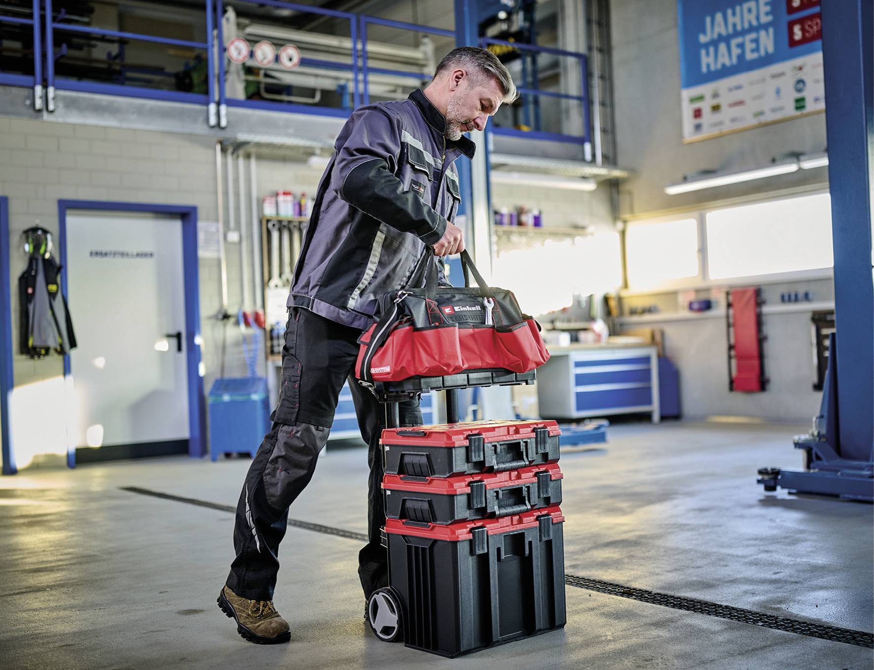 Muž v pracovním oblečení táhne červenou brašnu s nářadím přes dílnu s hydraulickými zvedáky a nástroji na pojízdném vozíku.