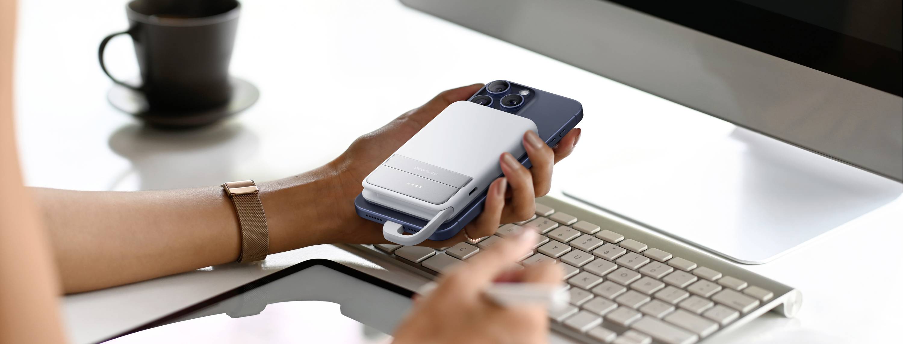 Osoba drží smartphone před monitorem počítače, v pozadí šálek. Scéna zobrazuje moderní práci v kanceláři.