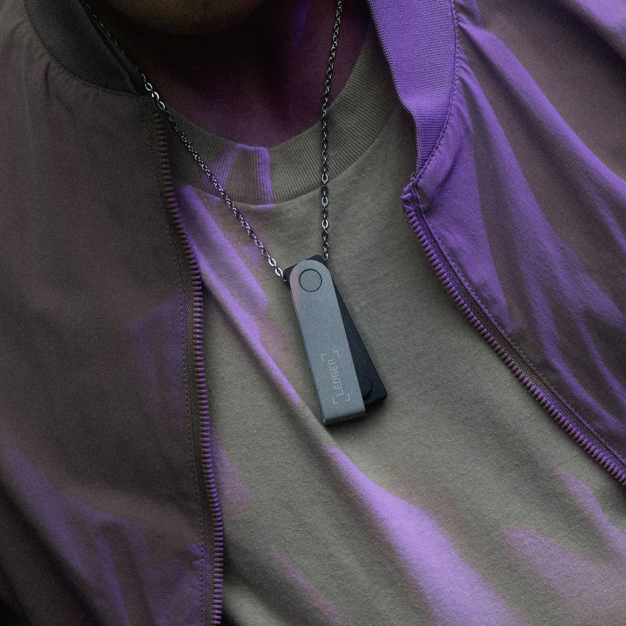 Osoba nosí náhrdelník s obdélníkovým USB diskem jako přívěskem, před fialovým svetrem a khaki tričkem.