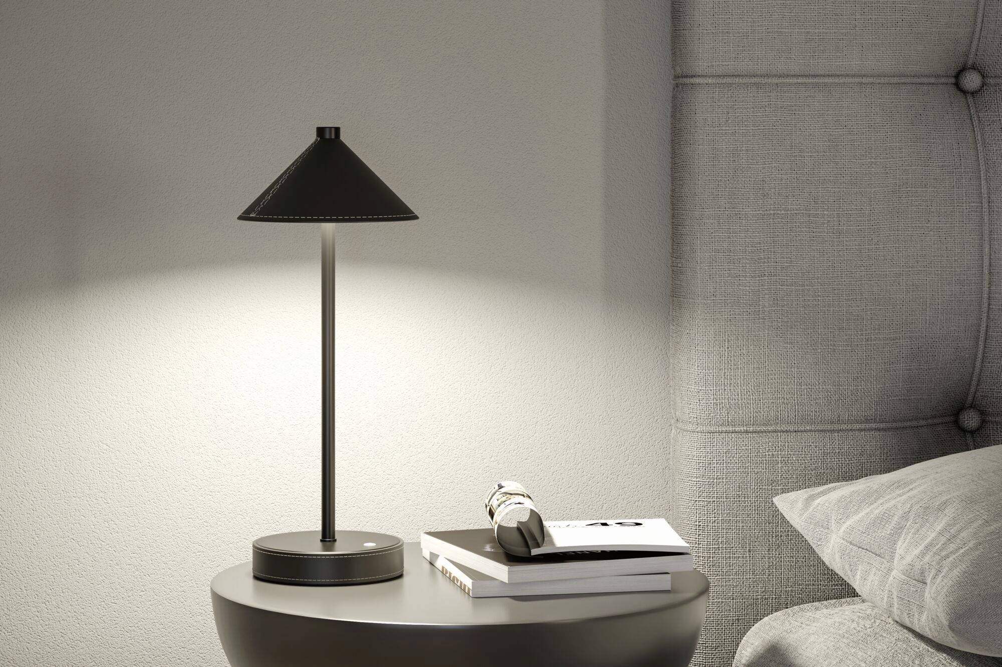 Stolní lampa s moderním designem stojí na kulatém nočním stolku. Vedle lampy leží knihy a brýle.