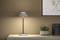 Moderní stolní lampa svítí na světlém přisazeném stolku vedle rostliny. Na stěně visí minimalistické umělecké dílo.