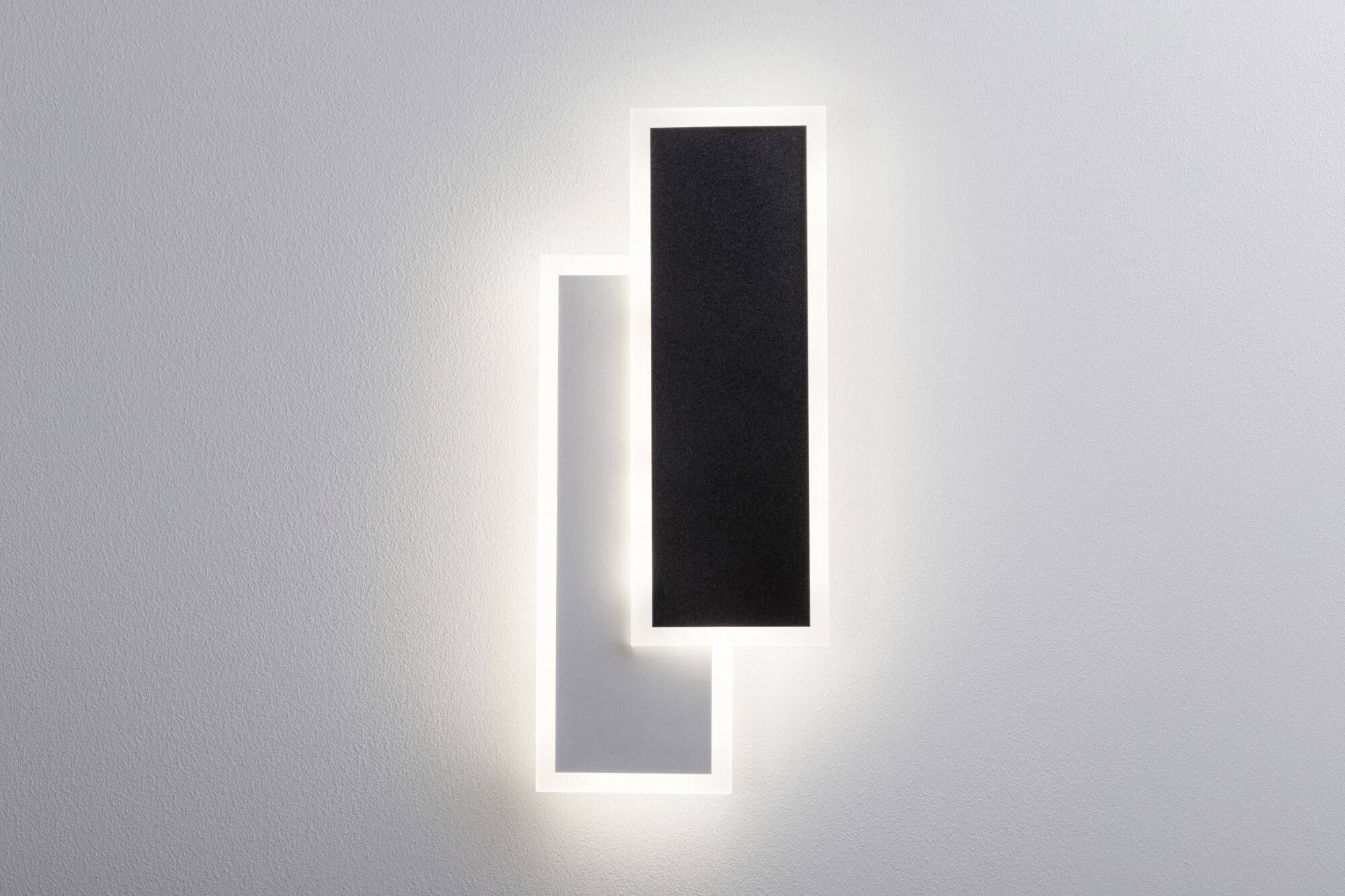 Nástěnná lampa se dvěma překrývajícími se obdélníkovými panely, jeden černý, jeden bílý, s jemným osvětlovacím efektem.