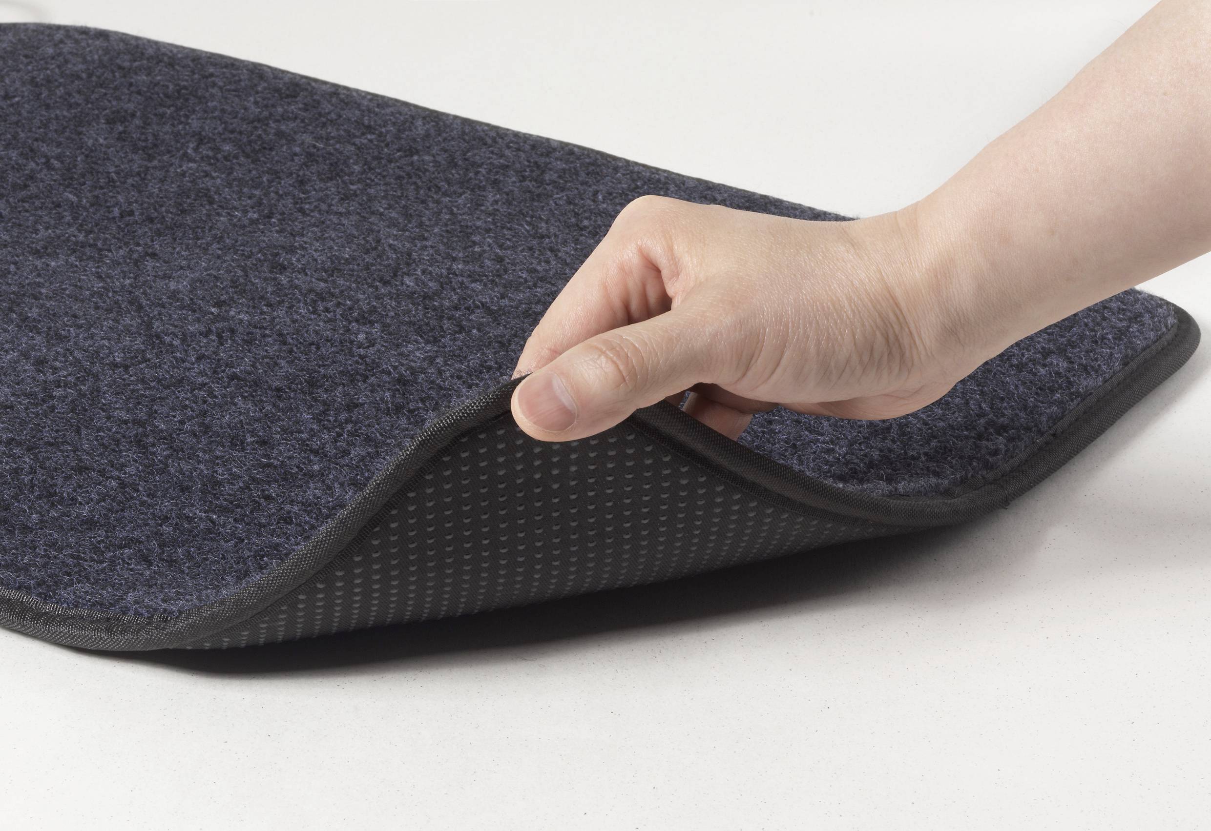 Ruka zvedá roh šedého rohožky a odhaluje protiskluzový povrch na spodní straně.