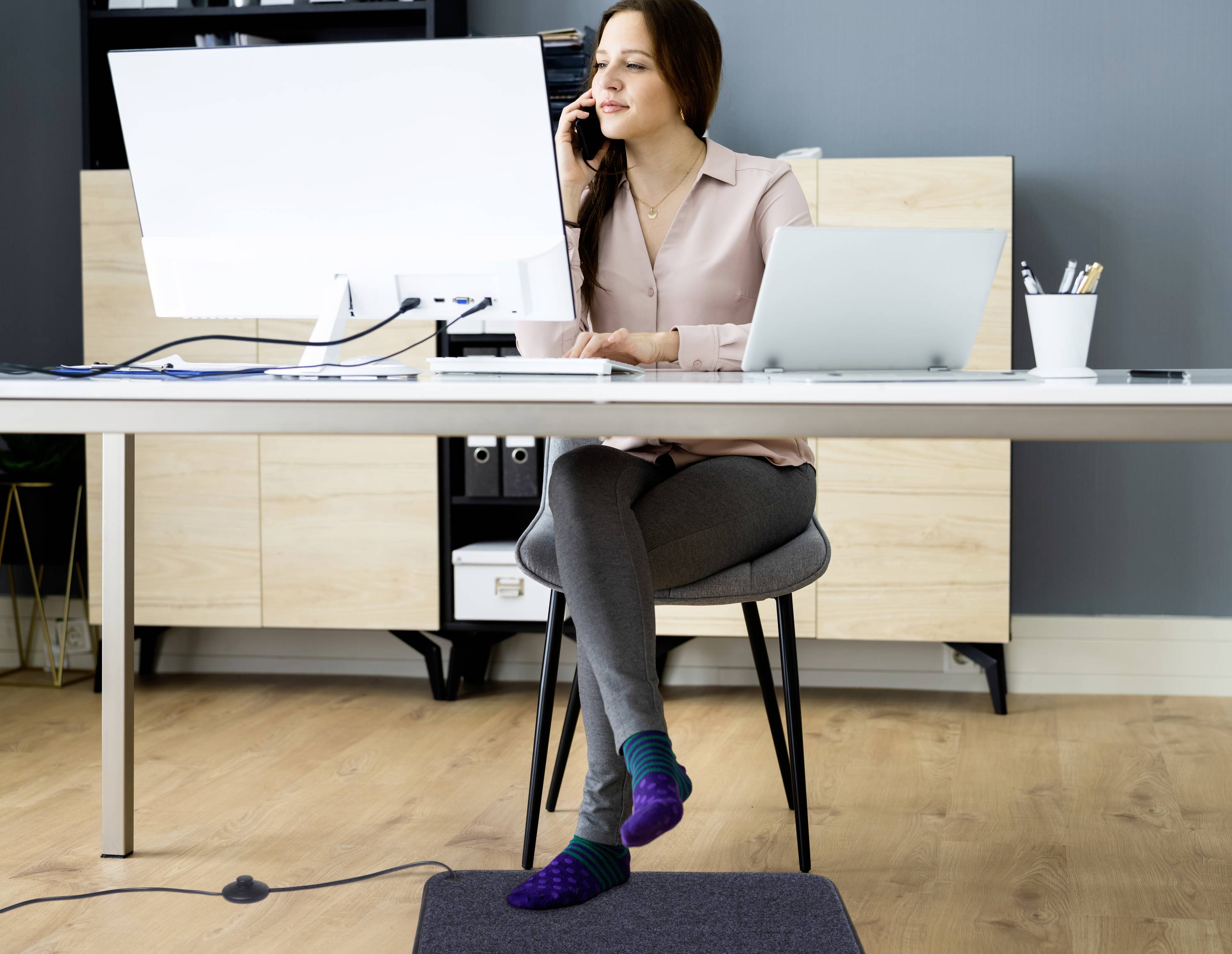 Žena sedí u stolu, telefon přitisknutý k uchu, a pracuje na počítači. Má ponožky a jednu nohu překříženou, působí soustředěným dojmem.