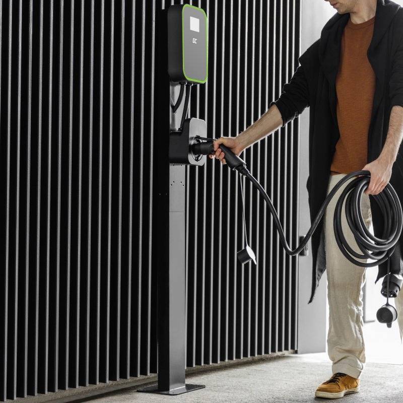Osoba v světlých kalhotách a tmavém plášti nabíjí elektromobil na dobíjecí stanici v moderním interiéru.