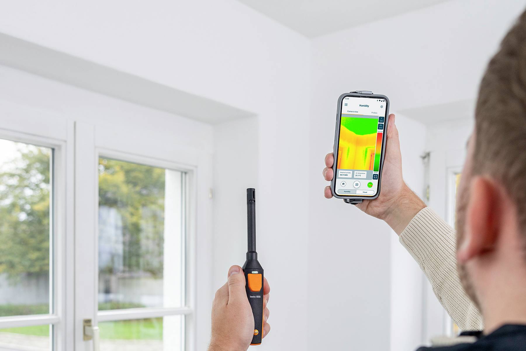 Jedna osoba používá digitální měřicí přístroj a chytrý telefon k analýze tepelných ztrát u okna. Na obrazovce je zobrazena teplotní mapa.