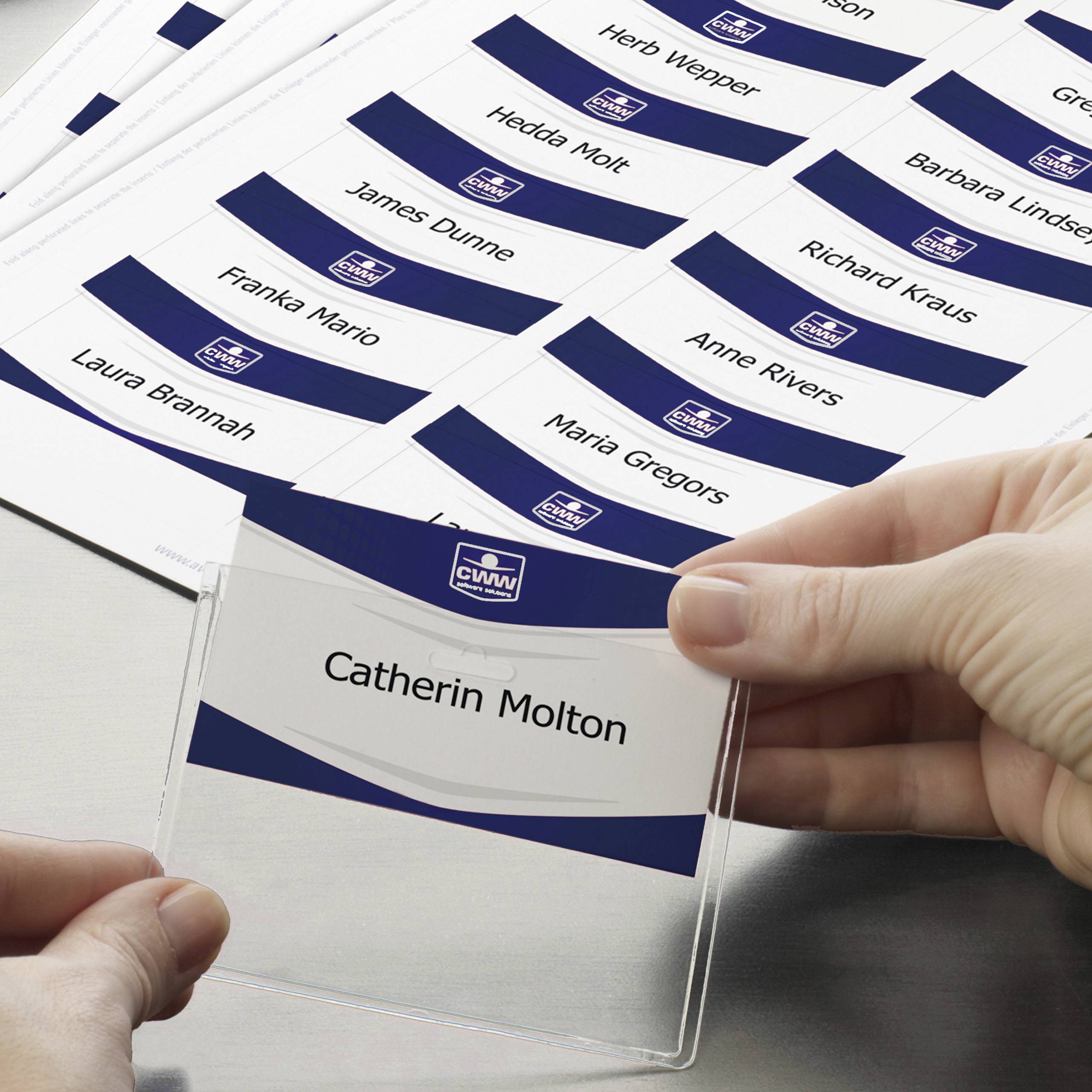 Ruka vkládá jmenovku s nápisem 'Catherine Molton' do držáku. V pozadí jsou další jmenovky.