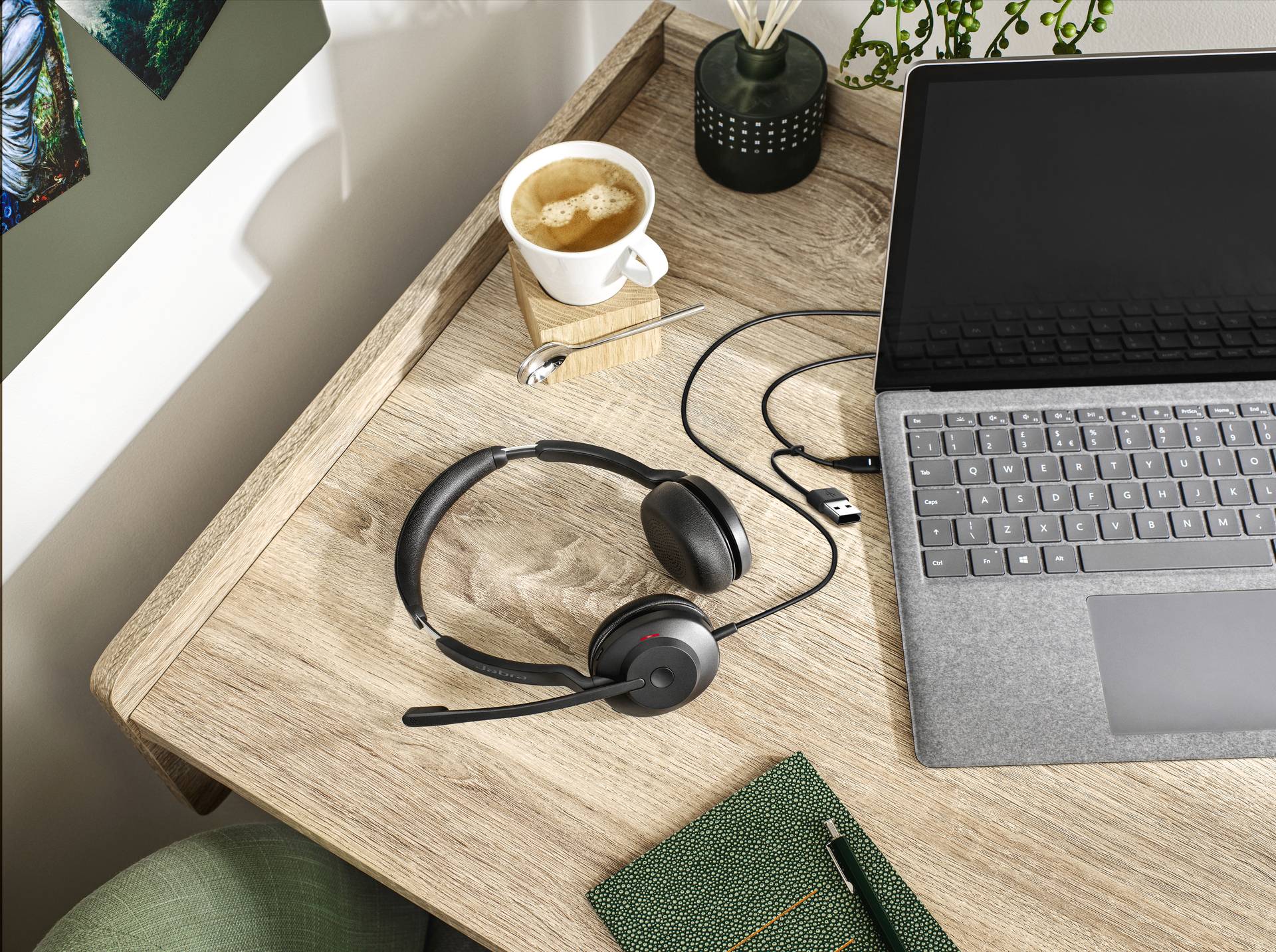 Laptop na dřevěném stole s připojenou sluchátkou, šálkem kávy a poznámkovým blokem vyobrazuje domácí pracovní nebo studijní prostředí.