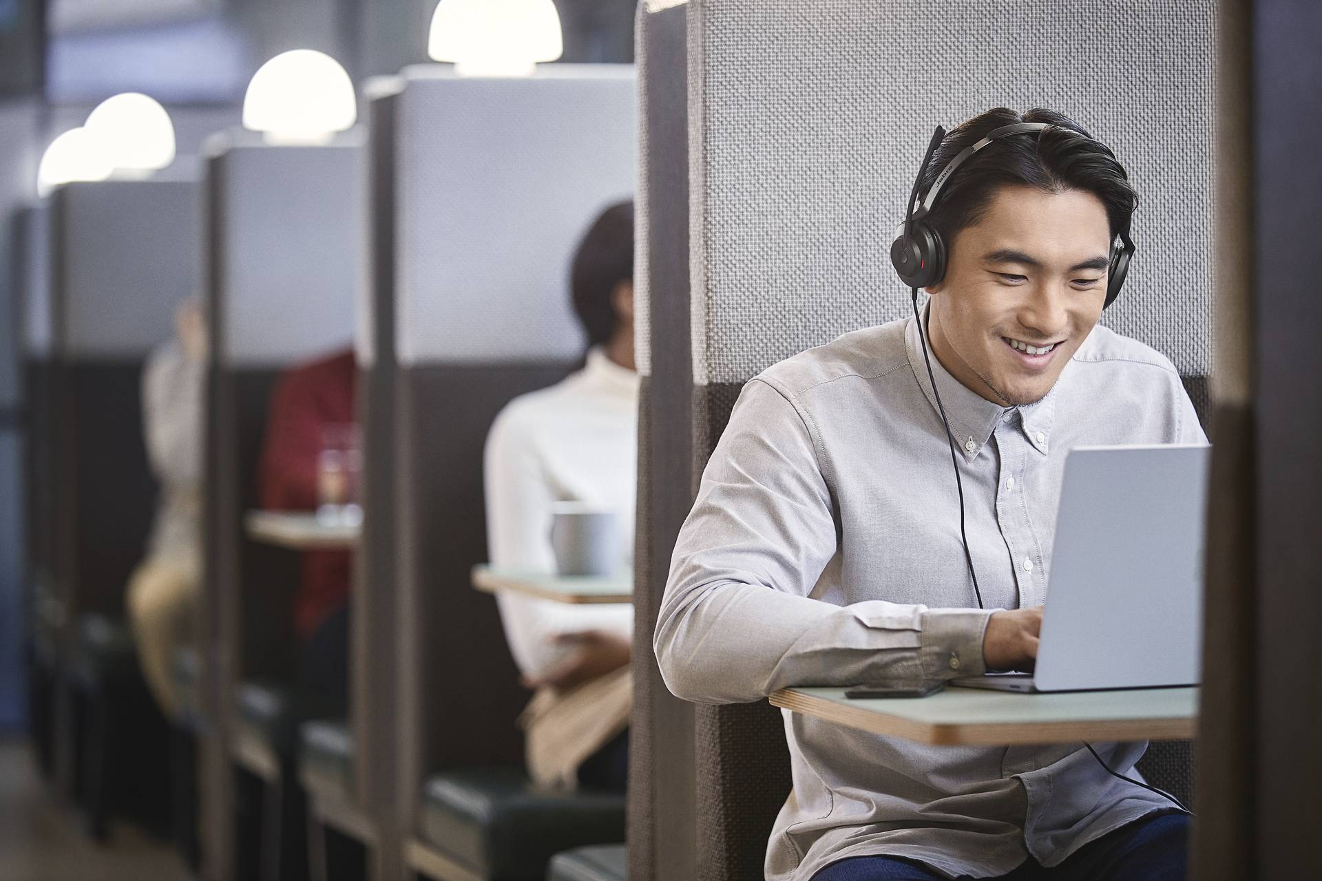 Muž se sluchátky, usmívající se, sedí u stolu v kavárně a pracuje na laptopu. V pozadí jsou vidět další lidé.