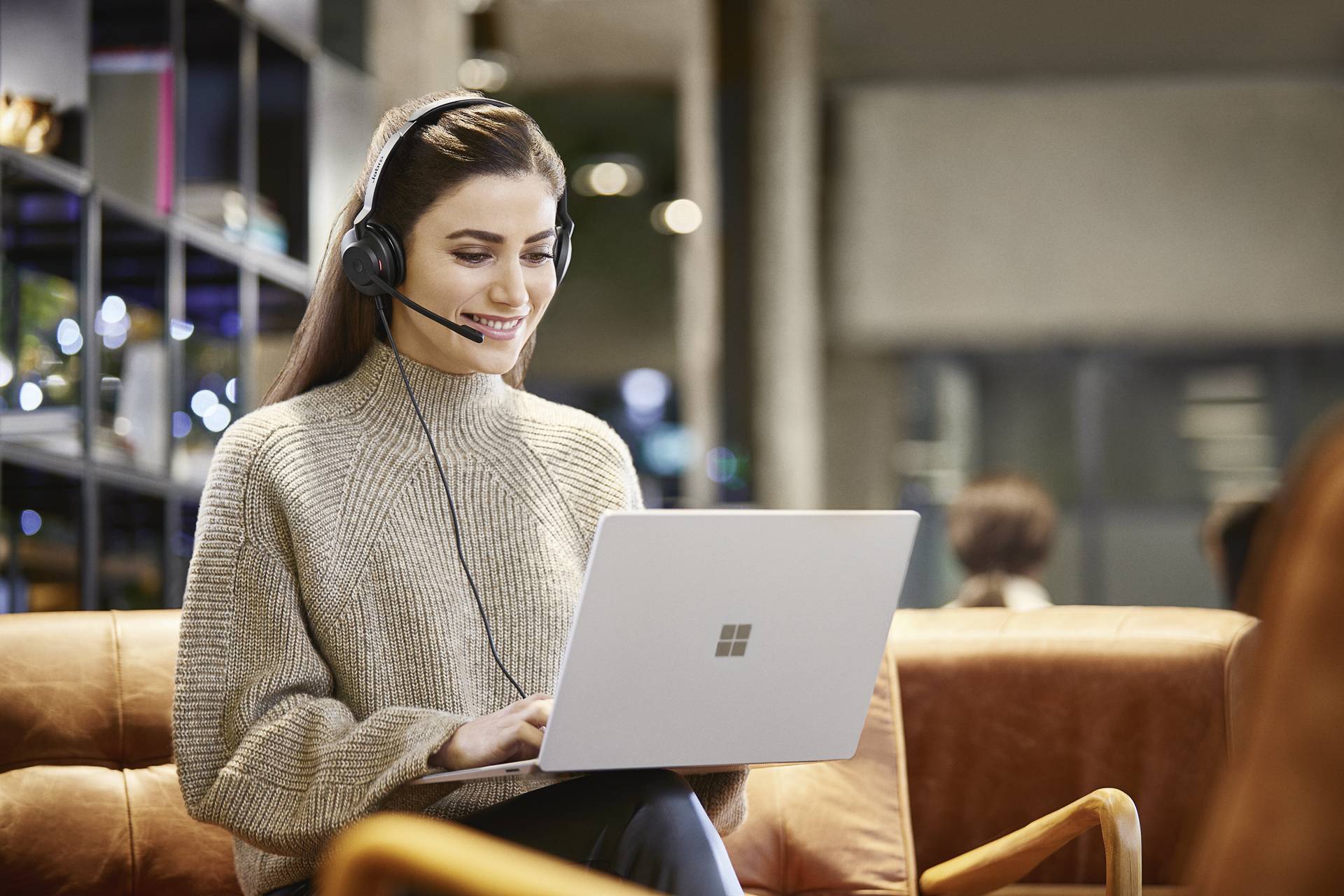 Žena s nasazenými sluchátky sedí na koženém gauči a pracuje na laptophu, přičemž se usmívá. Nachází se v moderním pracovním prostředí.