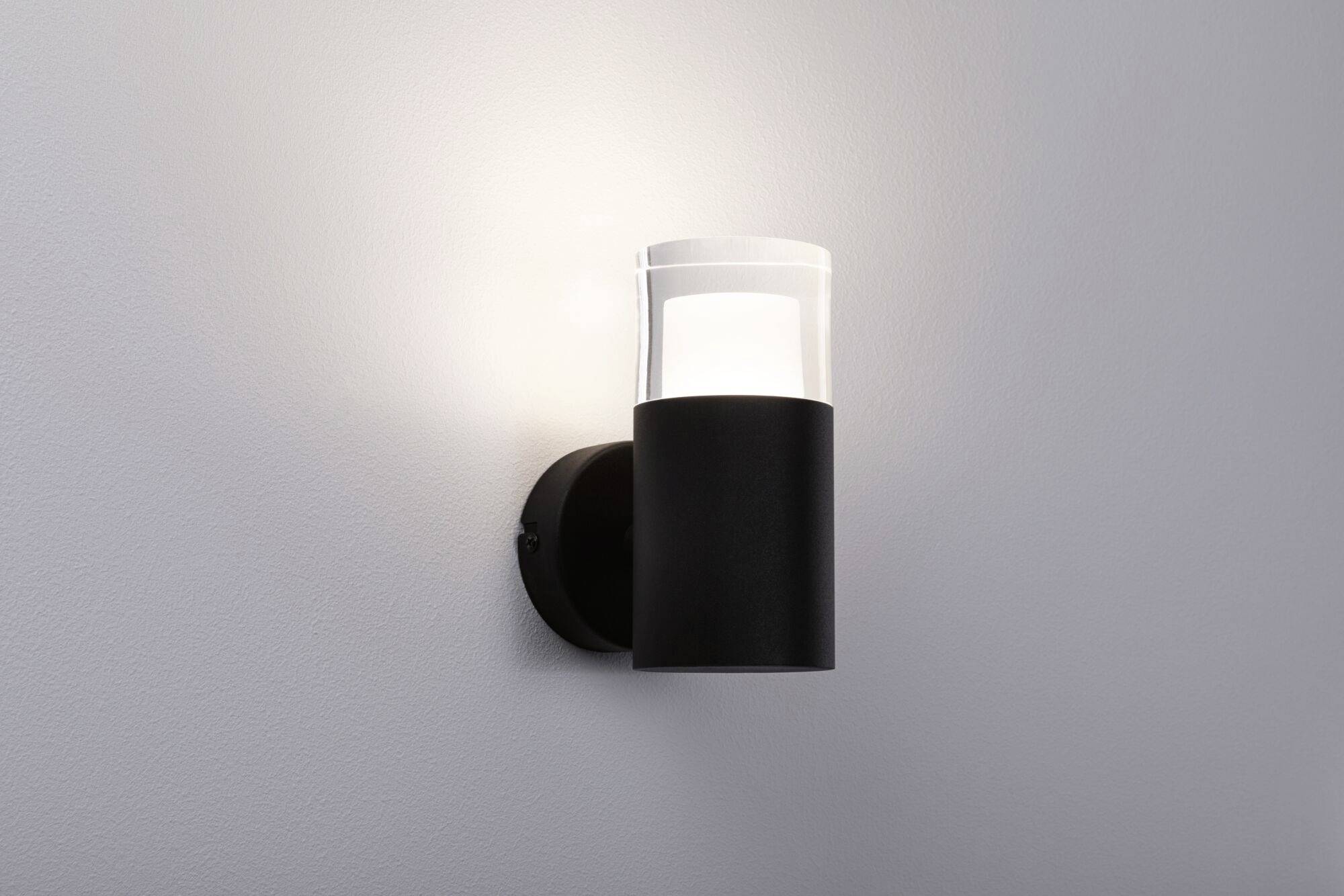 Nástěnná lampa s černou základnou a průhledným stínidlem, která svítí na bílé zdi a vytváří jemné osvětlení.