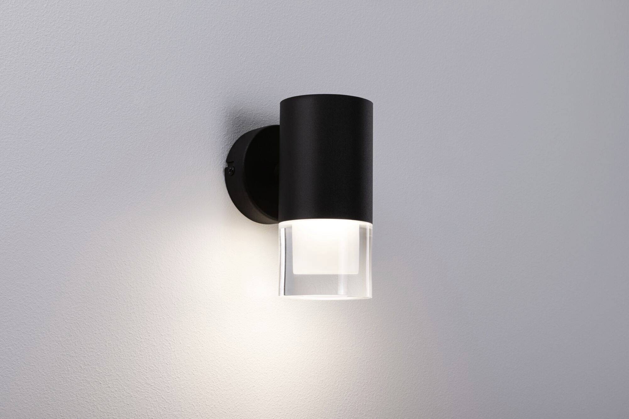 Černá nástěnná lampa svítí na světlé stěně, moderně a minimalisticky navržená, vyzařující teplé světlo dolů.