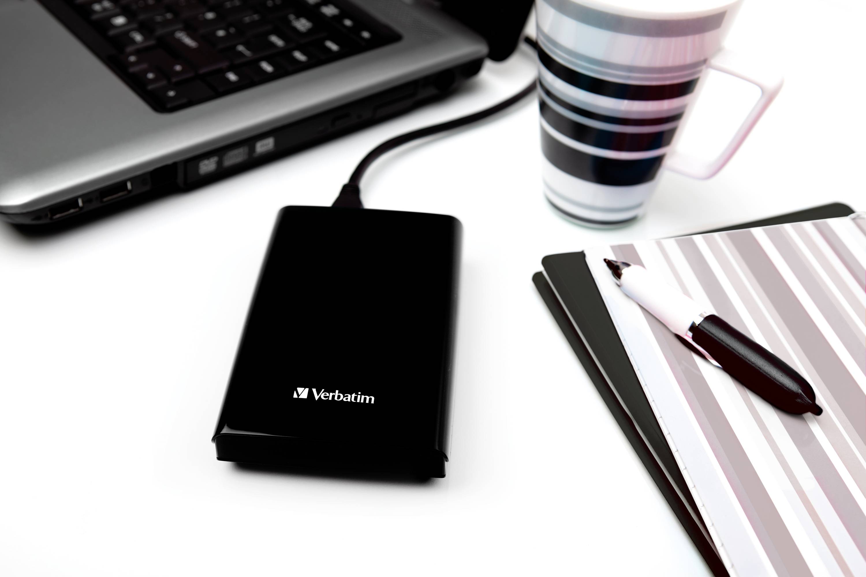 Externí pevný disk vedle laptopu, pruhované šálky a poznámkového bloku s perem na pracovním stole.