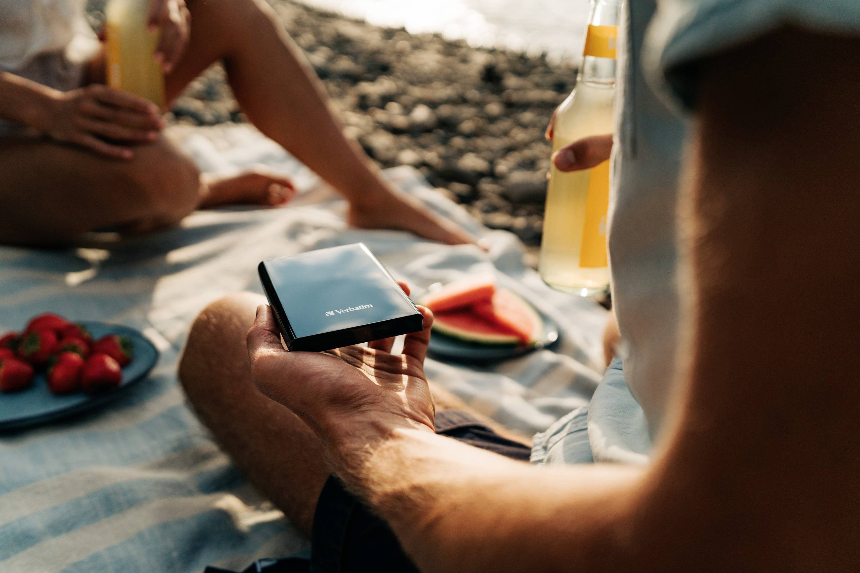 Osoba drží smartphone při pikniku na pláži. V pozadí sedí další lidé s nápoji a ovocem, jako jsou jahody a melouny.