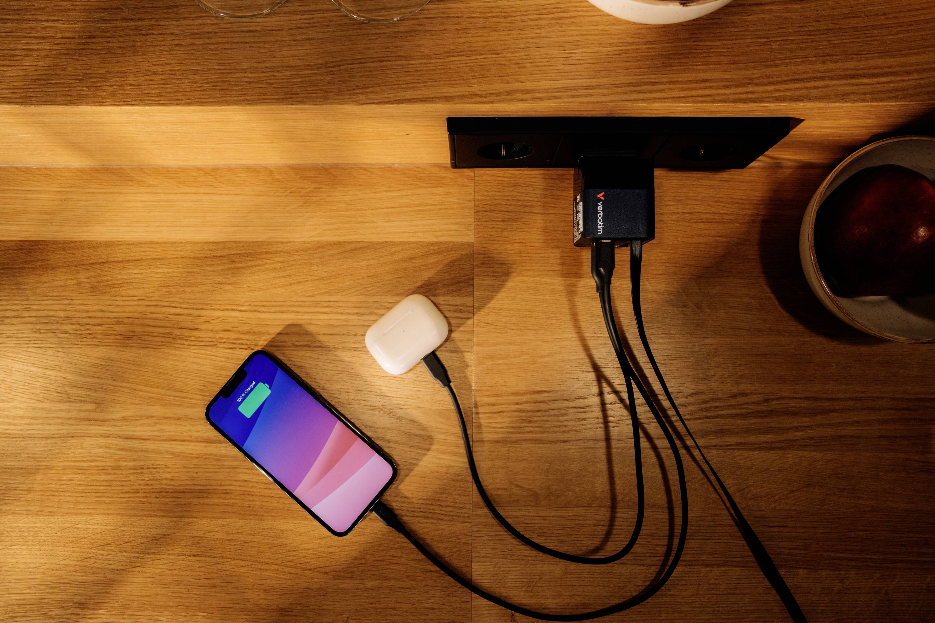 Smartphone a bezdrátová sluchátka jsou současně nabíjena pomocí nabíječky připojené ke zdi na dřevěném stole.