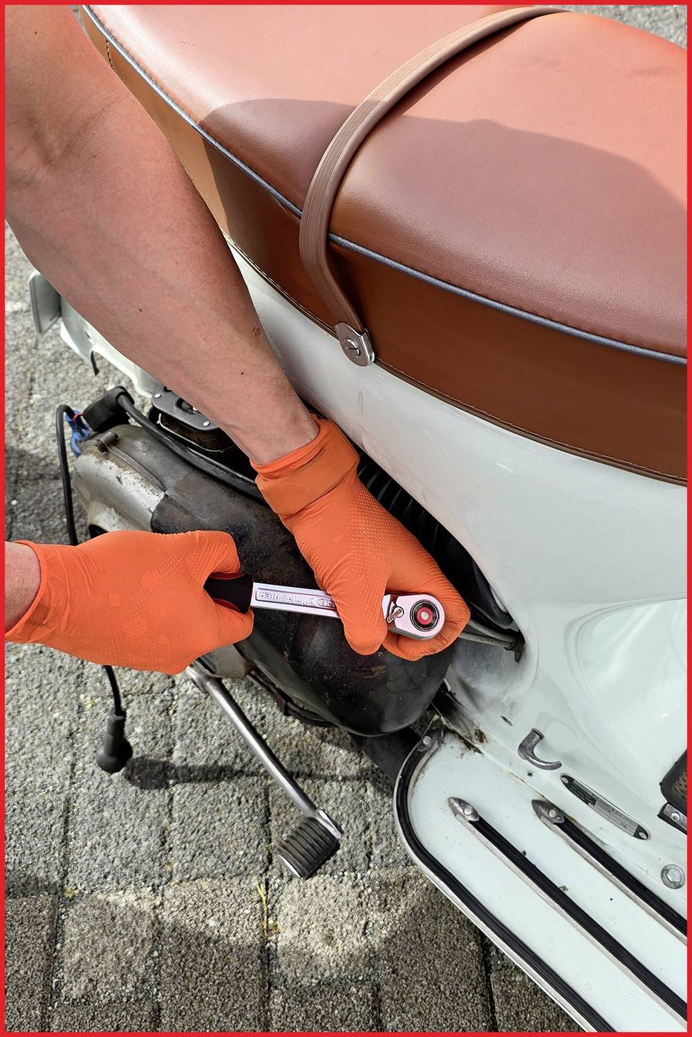 Osoba v oranžových rukavicích opravuje bílou motorku, používá klíč na motoru. Sedí na dlážděné ploše.